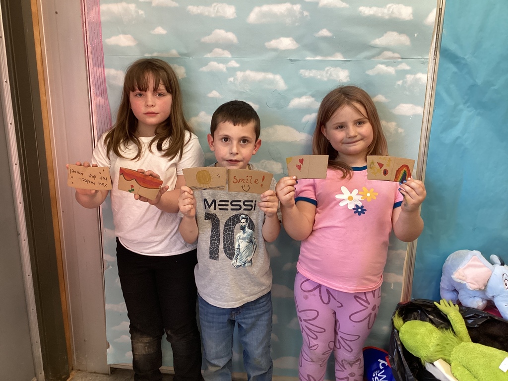 Three young students stand in front of a cloud-themed backdrop, holding decorated coffee sleeve cards with colorful drawings and messages of kindness, including “Smile!”