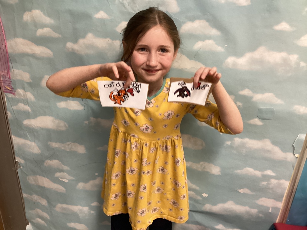 A student in a yellow dress holds up two small illustrated cards featuring hand-drawn animals and playful designs, smiling in front of a cloud-themed backdrop.