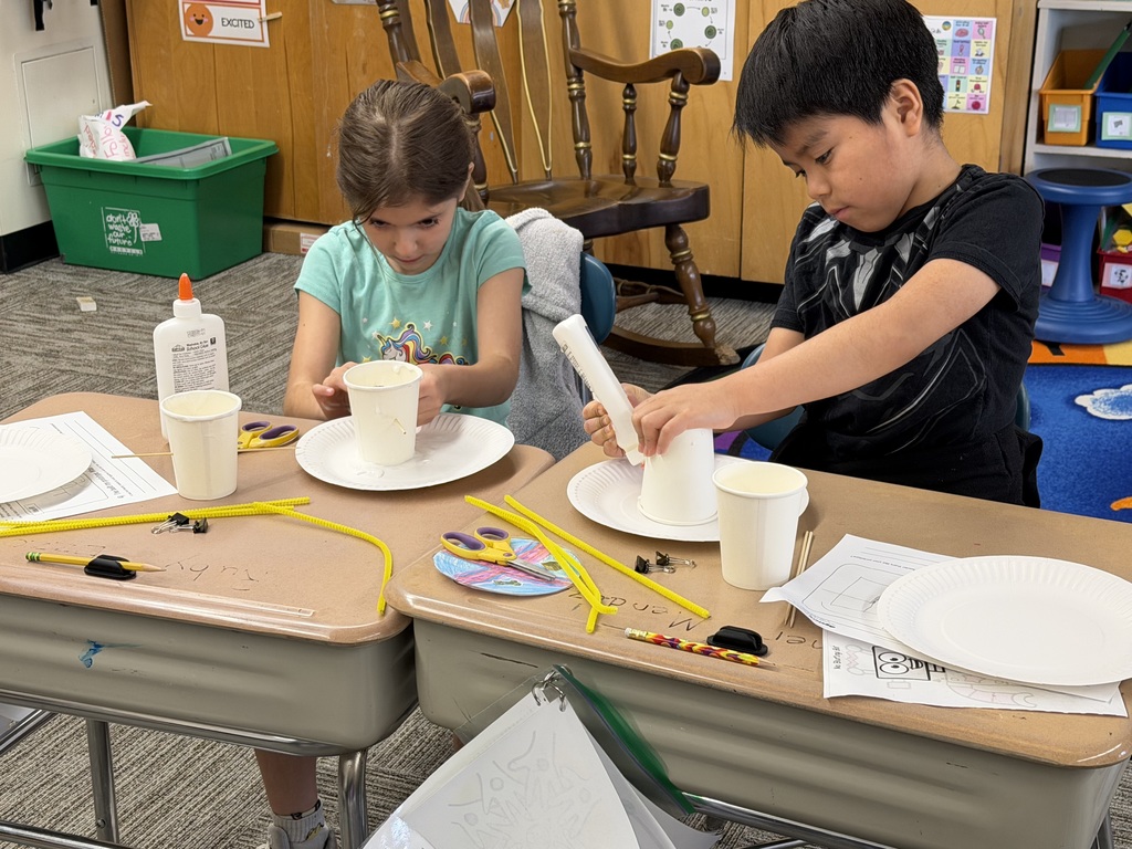 Two students concentrate on gluing and assembling birdhouse prototypes using paper cups and plates, with craft tools and materials arranged on their desks.