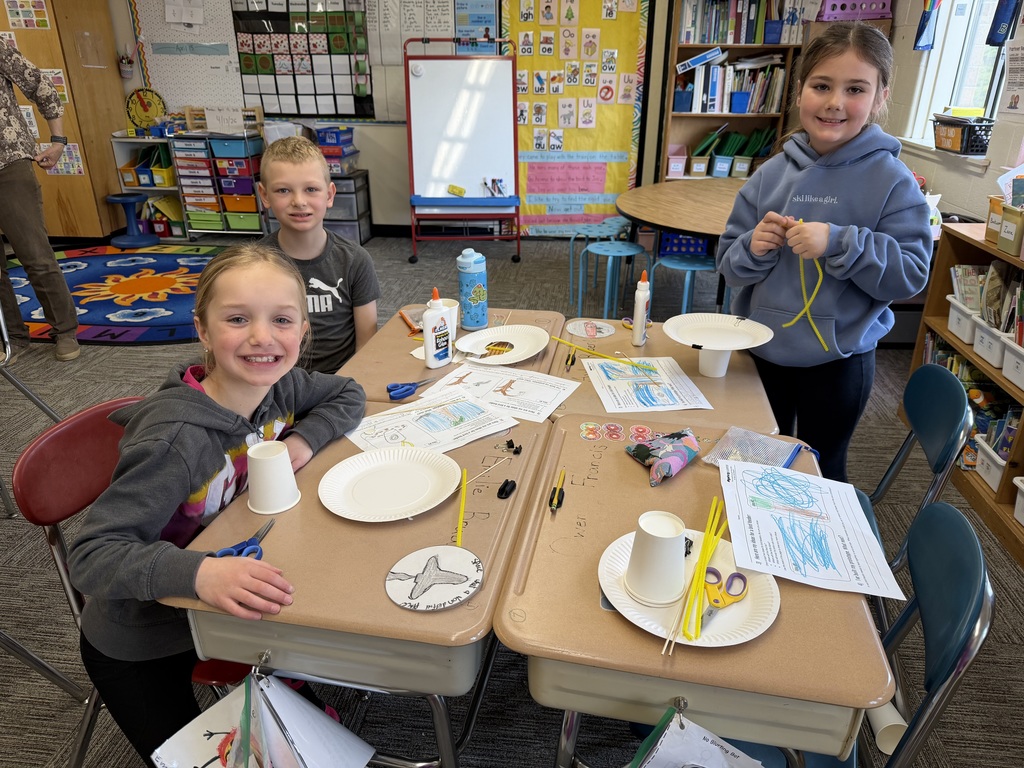 Three students sit at a classroom table smiling and working on birdhouse prototypes made from paper cups, plates, and craft materials, with scissors, glue, and worksheets spread out.
