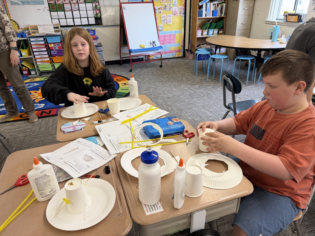 Two students work at desks creating birdhouse prototypes, carefully placing paper cups and materials together while following printed instructions.