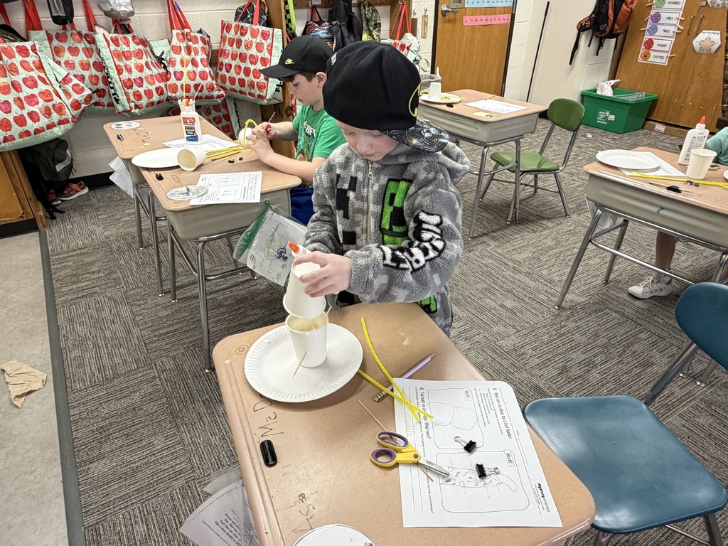 A student wearing a hat and jacket works independently at a desk, building a birdhouse prototype using paper cups, glue, and craft materials, with other students working in the background.