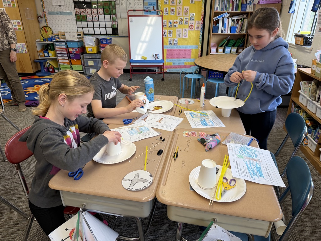 Three students focus on building their birdhouse prototypes, using glue and assembling paper cups and plates at a classroom table covered with supplies.