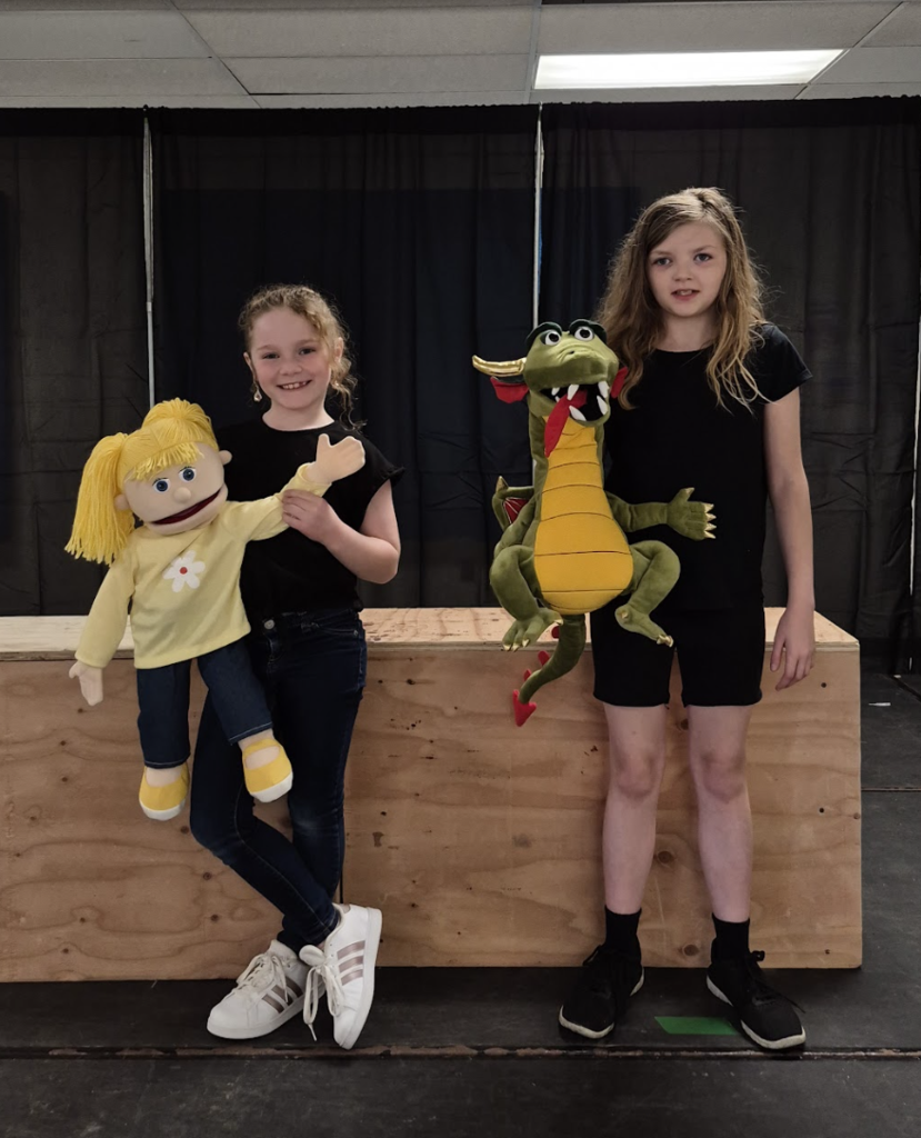 Two students stand on stage holding puppets—one a blonde girl puppet and the other a green dragon—posing in front of a black curtain backdrop.