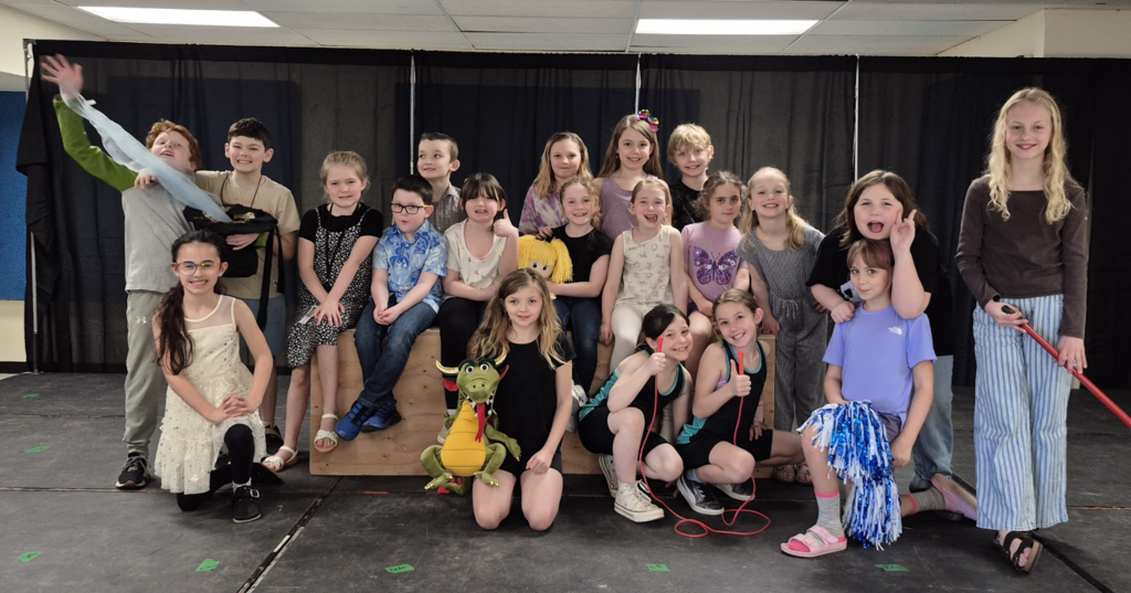 A group of students pose together on stage in casual clothes and costumes, smiling and gesturing while gathered around a wooden platform.