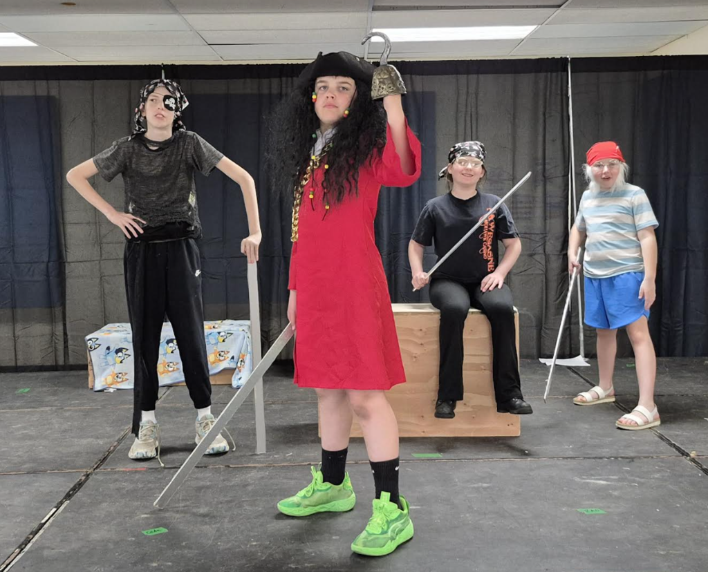 Four students dressed as pirates stand on stage holding prop swords, posing in front of a black curtain with a simple set piece nearby.