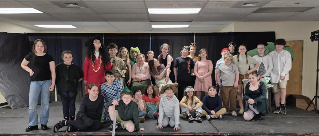 A large group of students gather across the stage in costumes and casual clothing, posing together in front of a black curtain during a group photo.