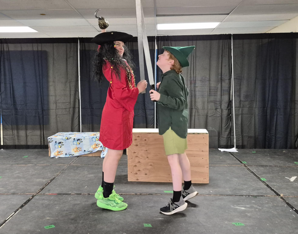 Two students face each other on stage holding prop swords, appearing to act out a scene while wearing costume pieces.