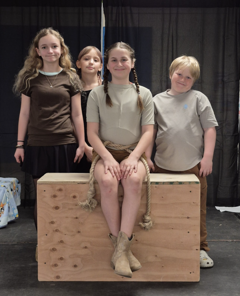 Four students pose together on stage, one seated on a wooden box with a rope prop, while others stand behind in costume-inspired outfits.