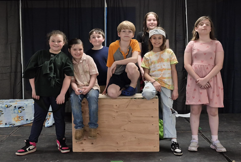 A group of students stand together on stage in casual and themed outfits, smiling and posing around a wooden platform.