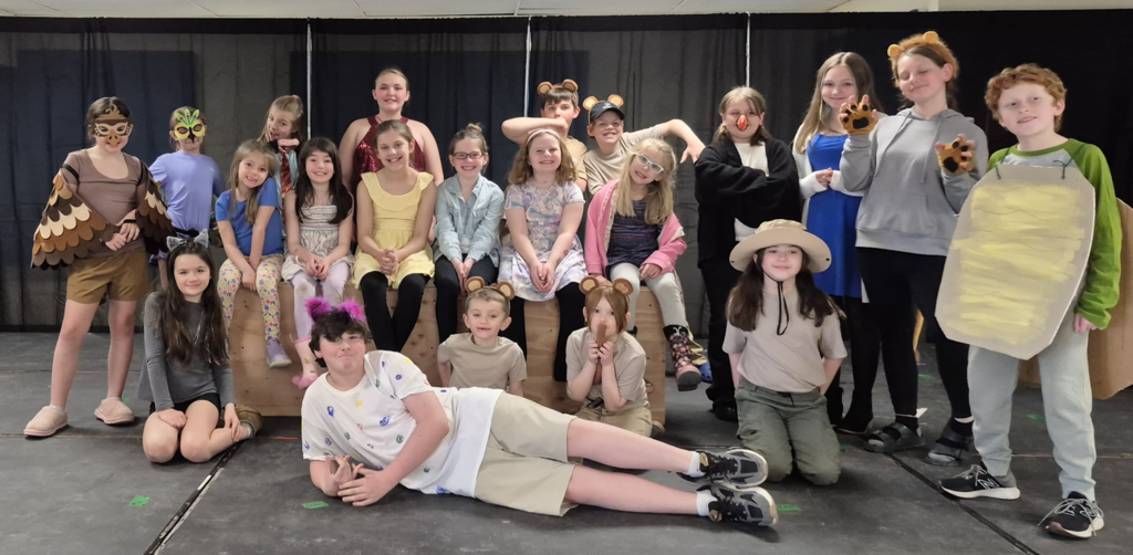 A large group of students pose on a stage in front of a black curtain, wearing a mix of costumes including animals and themed outfits, smiling and posing together.