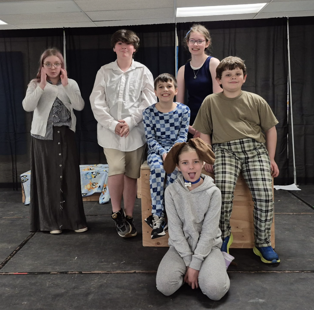 Six students pose on stage in character costumes, including casual and themed outfits, standing and sitting around a wooden box.