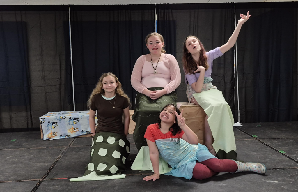 Four students pose on stage in mermaid-inspired costumes, sitting and kneeling while smiling and gesturing playfully.