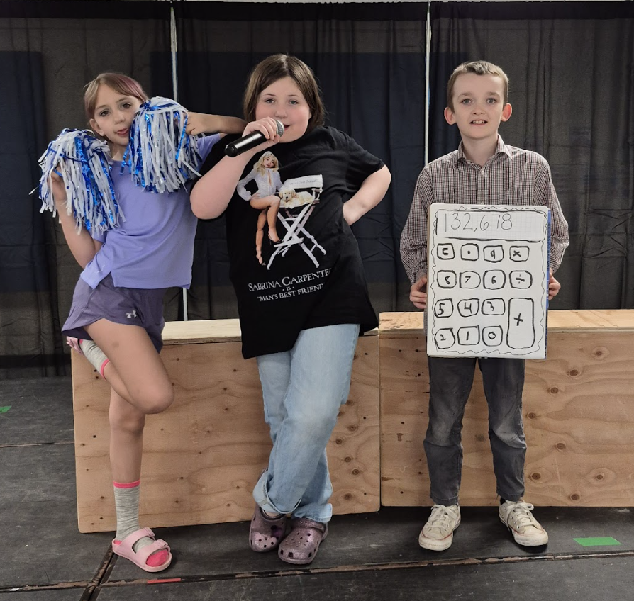Three students pose on stage, one holding pom-poms, one singing into a microphone, and another wearing a handmade calculator costume.