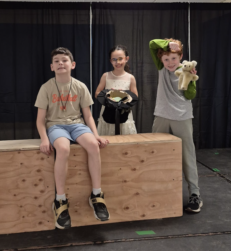 Three students pose on stage with props, including a top hat and a stuffed animal, smiling and gesturing playfully.