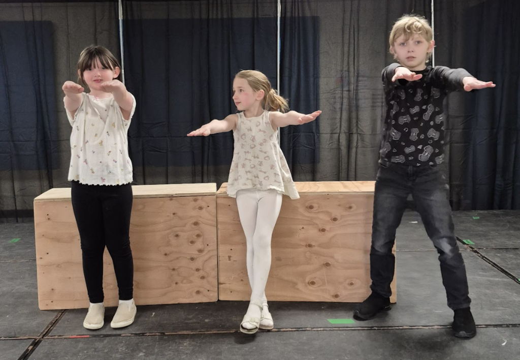 Three students stand in a row on stage with arms extended forward, as if mid-performance or dance pose.