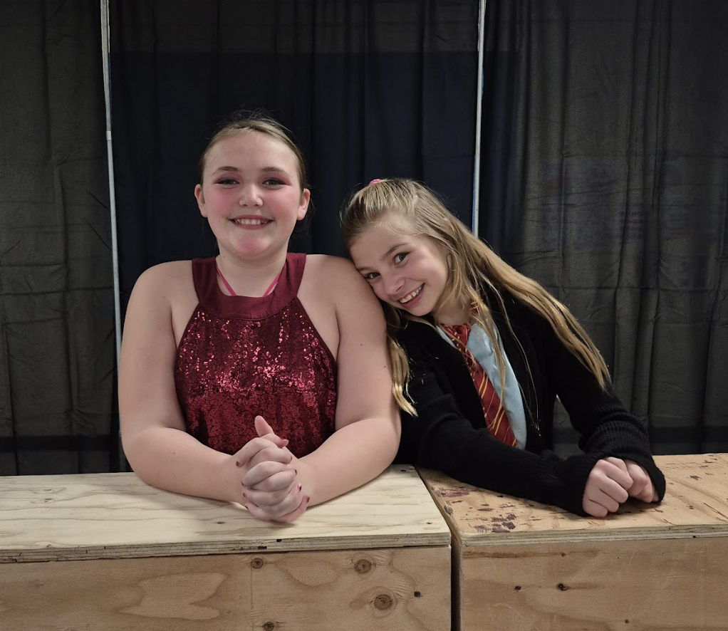 Two students lean on wooden stage blocks, smiling at the camera—one in a red sequined top, the other in a costume with a striped tie and cardigan.