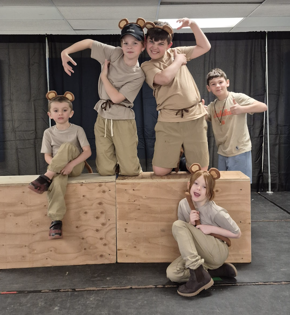 Five students dressed in tan outfits with bear ears pose on stage, some crouching and others flexing playfully.
