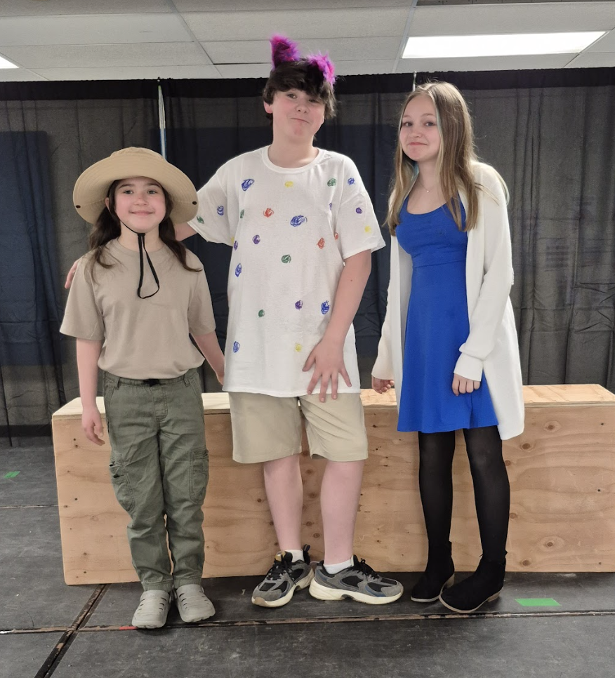 Three students pose together on stage in mixed costumes, including a safari-style outfit, a colorful shirt with cat ears, and a blue dress.