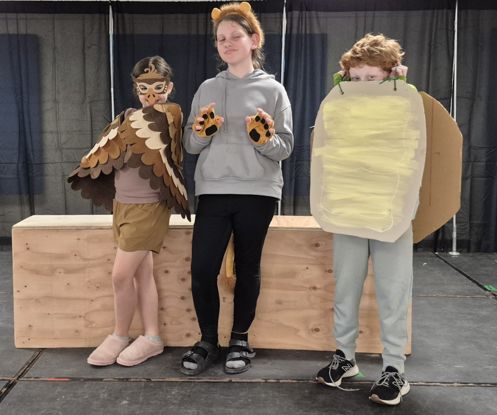 Three students pose in animal-themed costumes, including an owl outfit, bear ears and paws, and a large cardboard costume piece.