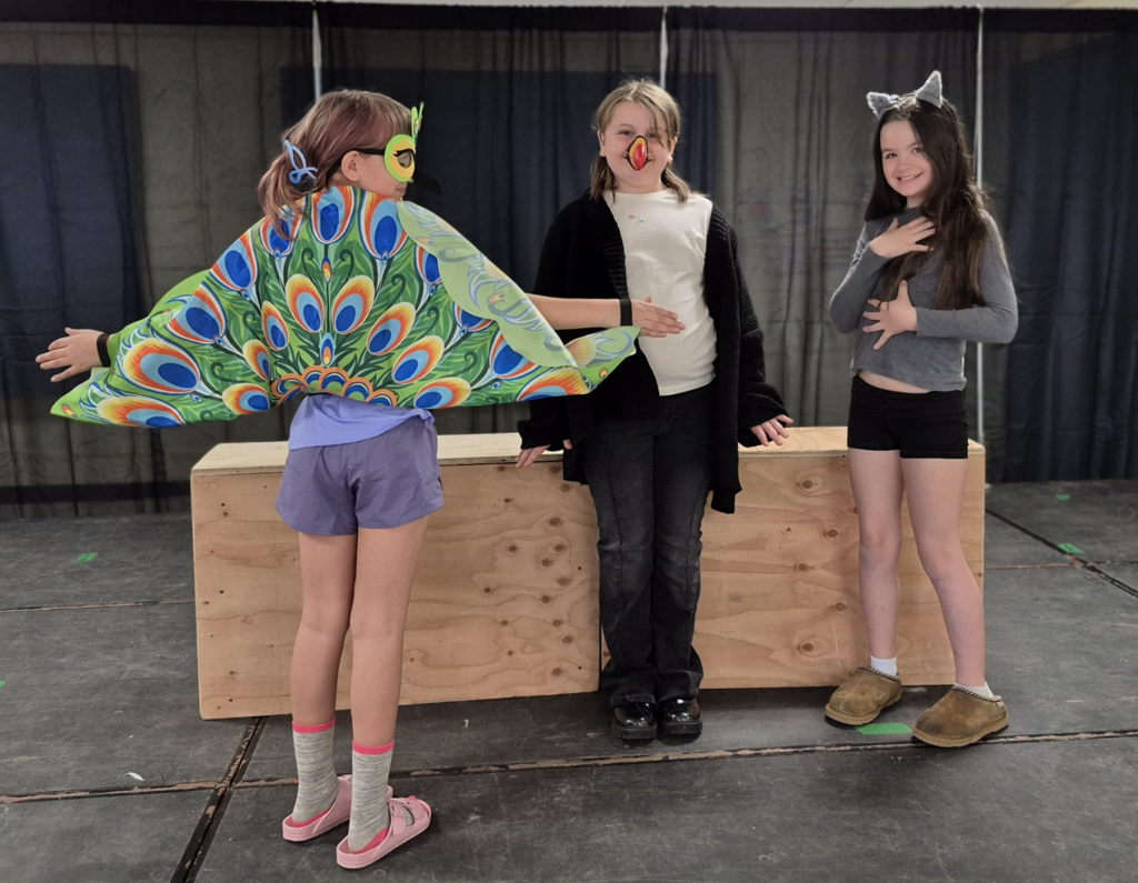 Three students pose on stage, one wearing a bright peacock cape with wings spread, while the others wear animal-inspired accessories.