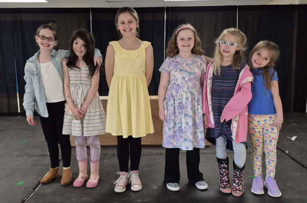 Six students stand side by side on stage in colorful dresses and casual outfits, smiling with arms around each other.