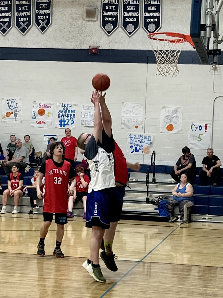 Two players jump near the basket attempting a shot, while others watch from the court and bleachers during a game.