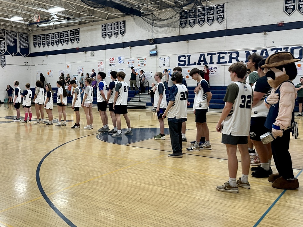 A line of students in basketball uniforms stands on the court during pregame, facing the flag with hands over their hearts while spectators watch from the bleachers.