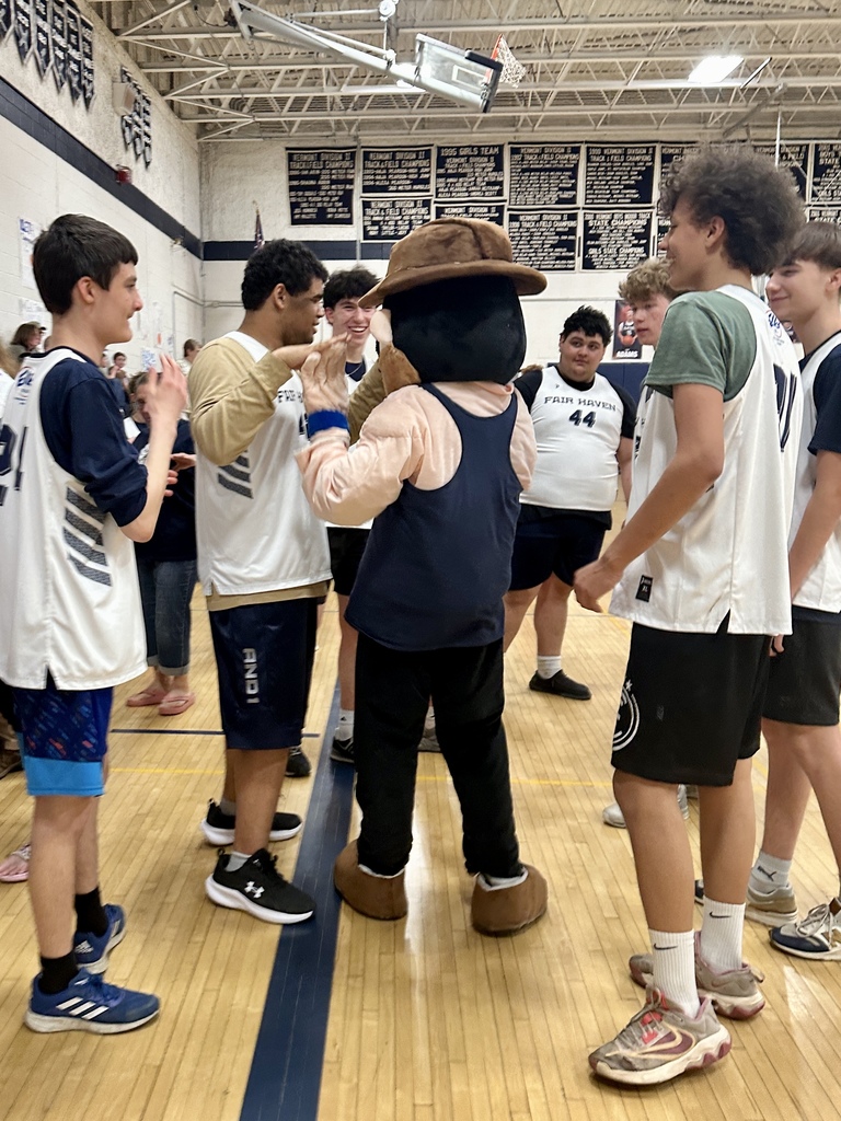Students in basketball uniforms gather in a circle in a gym, high-fiving and smiling around a school mascot in a hat.