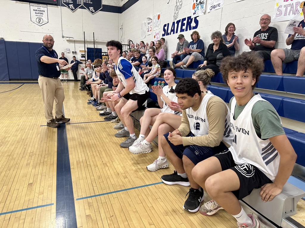 Basketball players sit on the bench clapping and cheering, while a coach stands nearby holding papers and addressing the team; spectators fill the bleachers behind them.
