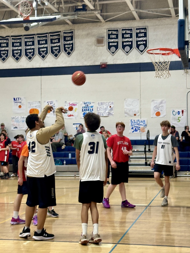 A player follows through on a jump shot as teammates and opponents watch the ball in the air, with handmade signs visible on the gym wall behind them.