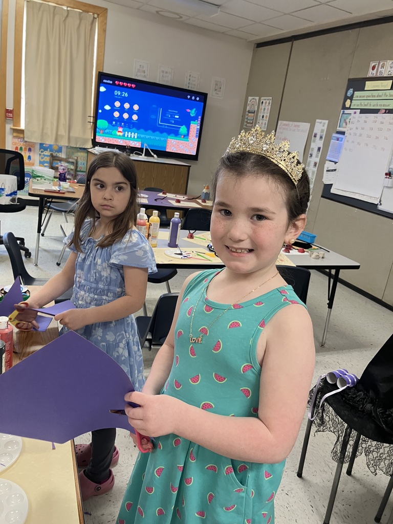   Two young girls working on an art project. The girl in the foreground smiles at the camera, wearing a teal dress with a watermelon print and a sparkling gold tiara. Behind her, another girl in a blue floral dress is focused on cutting a piece of purple construction paper. A television in the background displays a colorful video game interface.