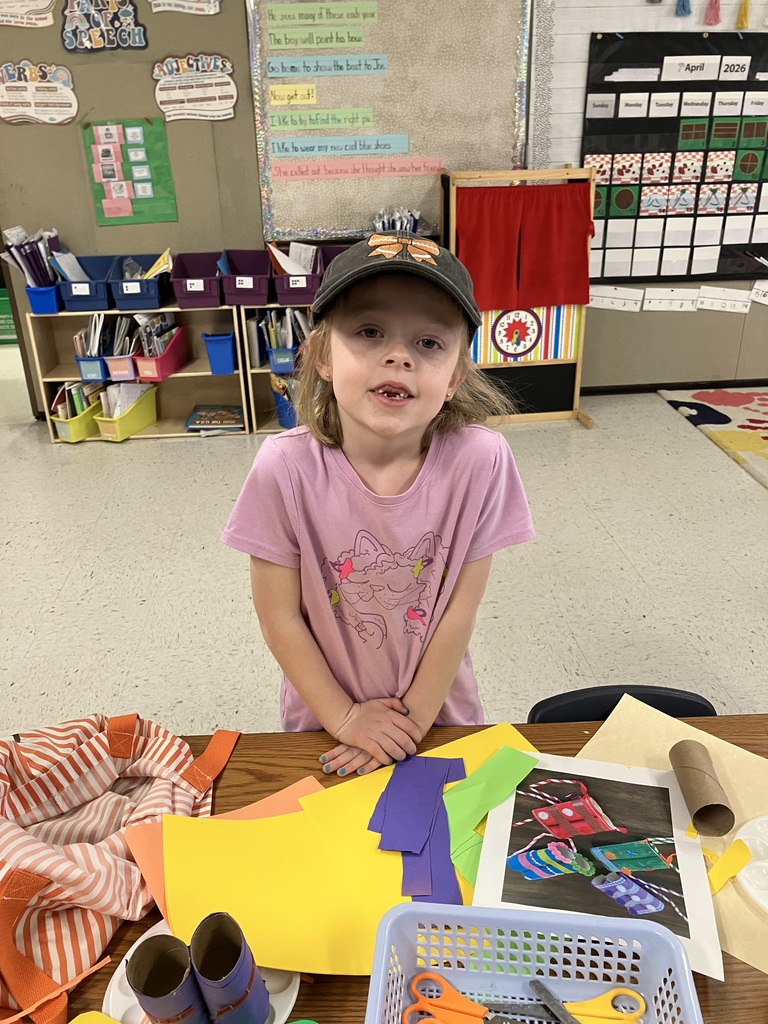   A medium shot of a young girl with a missing front tooth wearing a pink t-shirt and a dark baseball cap with a bow. She is standing behind a wooden table cluttered with colorful construction paper, scissors in a blue basket, and a reference photo of a craft project involving decorated cardboard tubes. 