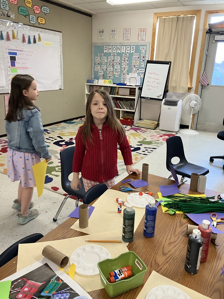 a young girl in a red cable-knit sweater stands at a craft table covered in supplies like paint, glue, and cardboard tubes. Another girl in a denim jacket and floral dress walks nearby. In the background, there is a colorful "handprint" rug and a white board that reads, "Today is Friday, April 17th, 2026."