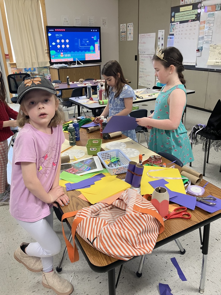  A medium shot of a young girl with a missing front tooth wearing a pink t-shirt and a dark baseball cap with a bow. She is standing behind a wooden table cluttered with colorful construction paper, scissors in a blue basket, and a reference photo of a craft project involving decorated cardboard tubes. 