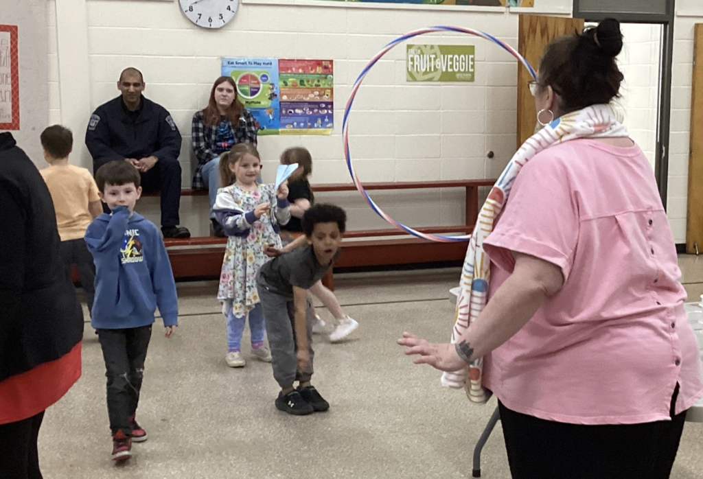 Students take turns throwing paper airplanes toward a hula hoop held by a staff member, while others watch and hold their own planes.