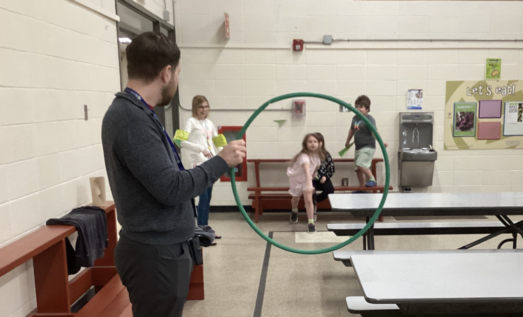 A teacher holds a green hula hoop while a student throws a paper airplane toward it, with other students waiting and watching in a cafeteria space.