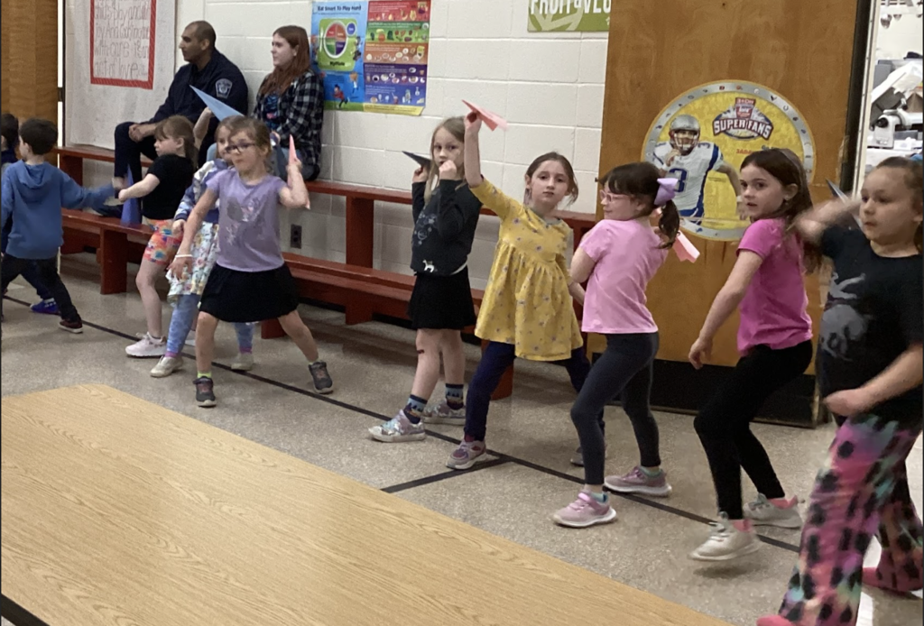A group of first grade students stands in a cafeteria line holding paper airplanes, preparing to throw them while adults sit on a bench behind them.
