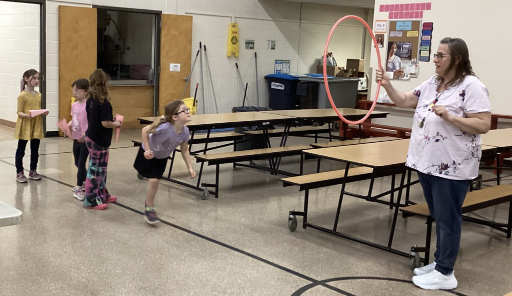 A student leans forward mid-throw, aiming a paper airplane toward a red hula hoop held by a teacher in a cafeteria with tables in the background.