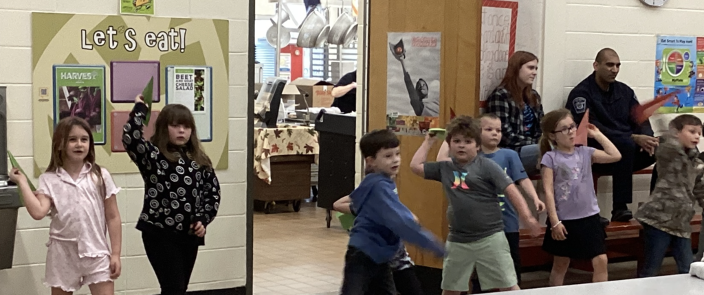 Young students line up with paper airplanes raised, preparing to throw, with a “Let’s eat!” poster and cafeteria serving area visible behind them.