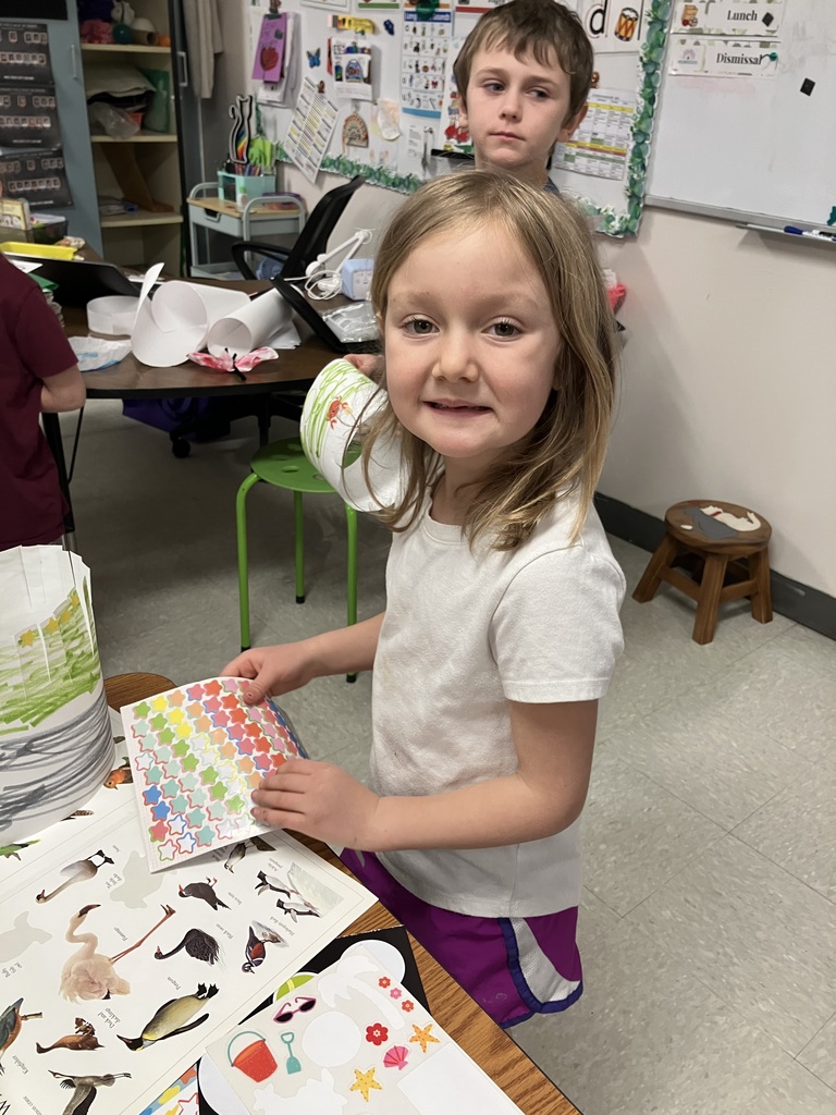 A close-up of a young girl in a white t-shirt holding a sheet of colorful star stickers. On the table in front of her is a white paper crown decorated with green grass and a crab sticker, along with a sheet of bird stickers. 