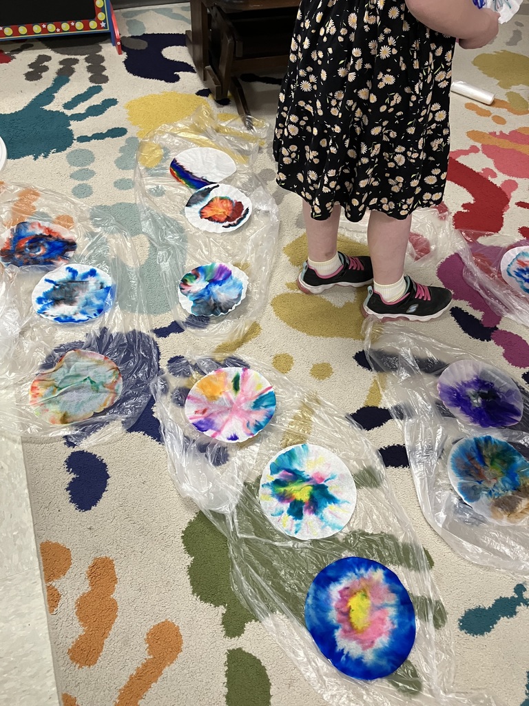  A high-angle shot of several coffee filters spread out on plastic sheets over a classroom rug. The filters show vibrant, bleeding patterns of blue, purple, yellow, and red ink. The legs of a child in a floral dress stand among the art.  Paper Crafting & Drawing 
