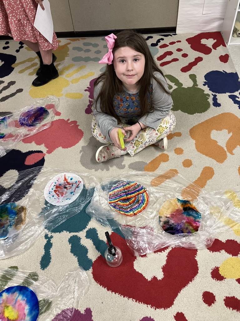 A young girl with a pink bow in her hair sits cross-legged on a colorful "handprint" patterned rug. In front of her are several coffee filters being decorated with colorful markers and a small spray bottle used to create a tie-dye effect.  