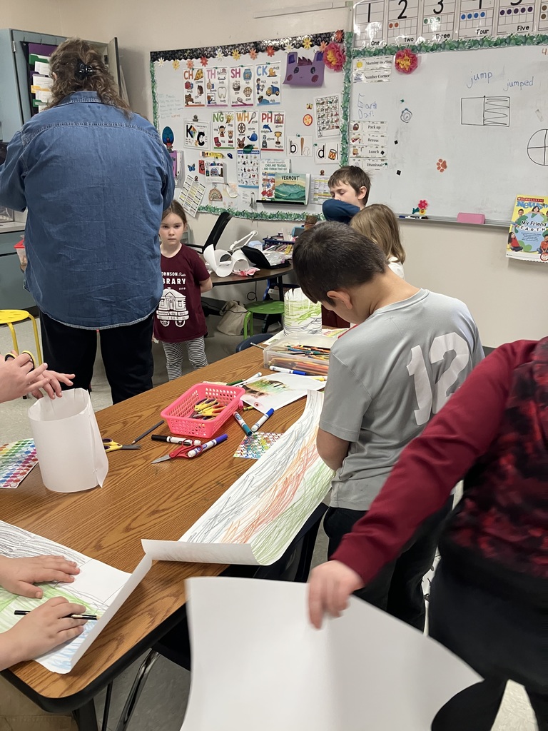A wide view of a classroom where children are working on various art projects. One child is drawing on a long strip of paper on the floor, while others work at wooden tables with supplies like red cups, pencils, and scissors.  