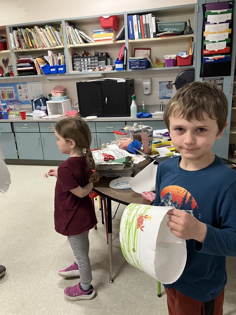  A boy in a blue long-sleeve shirt holds up a finished white paper crown featuring green scribbles and a crab sticker. In the background, another student stands near a shelf filled with books and colorful storage bins.