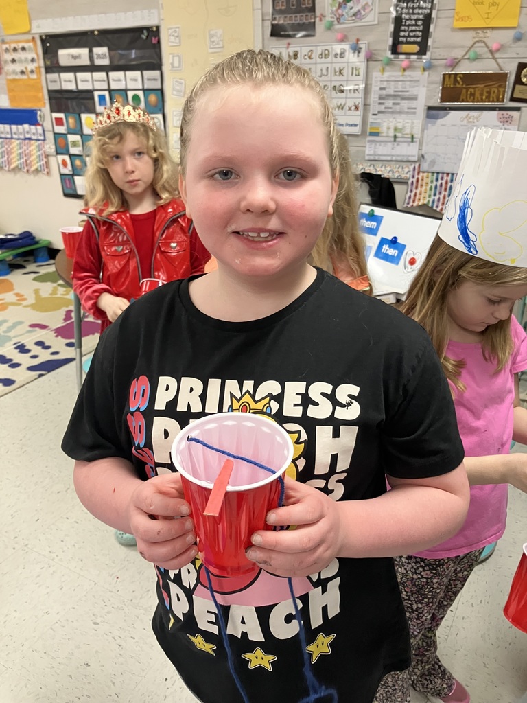 A young girl in a black "Princess Peach" T-shirt holds a red plastic cup with blue yarn and a wooden stick inside. She is smiling at the camera, and another girl in a red vest is visible in the background. 