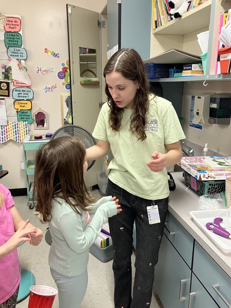  A teacher in a light green T-shirt leans down to talk to a young girl in a classroom. They are standing near a counter where a "Candy Land" board game and a hot glue gun are visible.