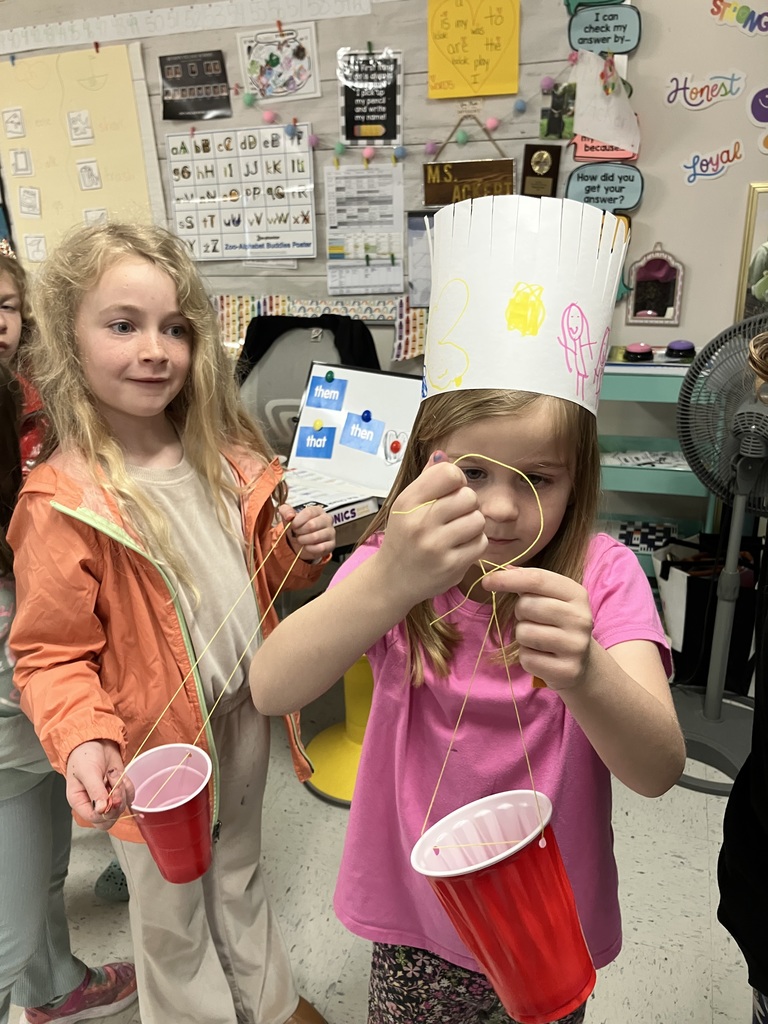 Two young girls work on their crafts. One girl in a pink shirt and a paper crown focuses on threading yellow string, while the girl next to her in an orange jacket holds her completed red cup craft. 