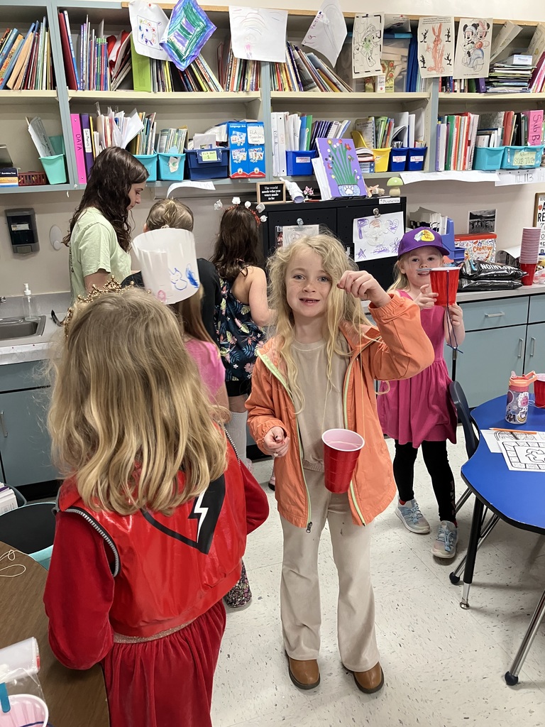 A wide shot of a classroom where several children are showing off their "cup and string" crafts. A girl in an orange jacket smiles at the camera, while another girl in a purple hat holds up her red cup in the background.  
