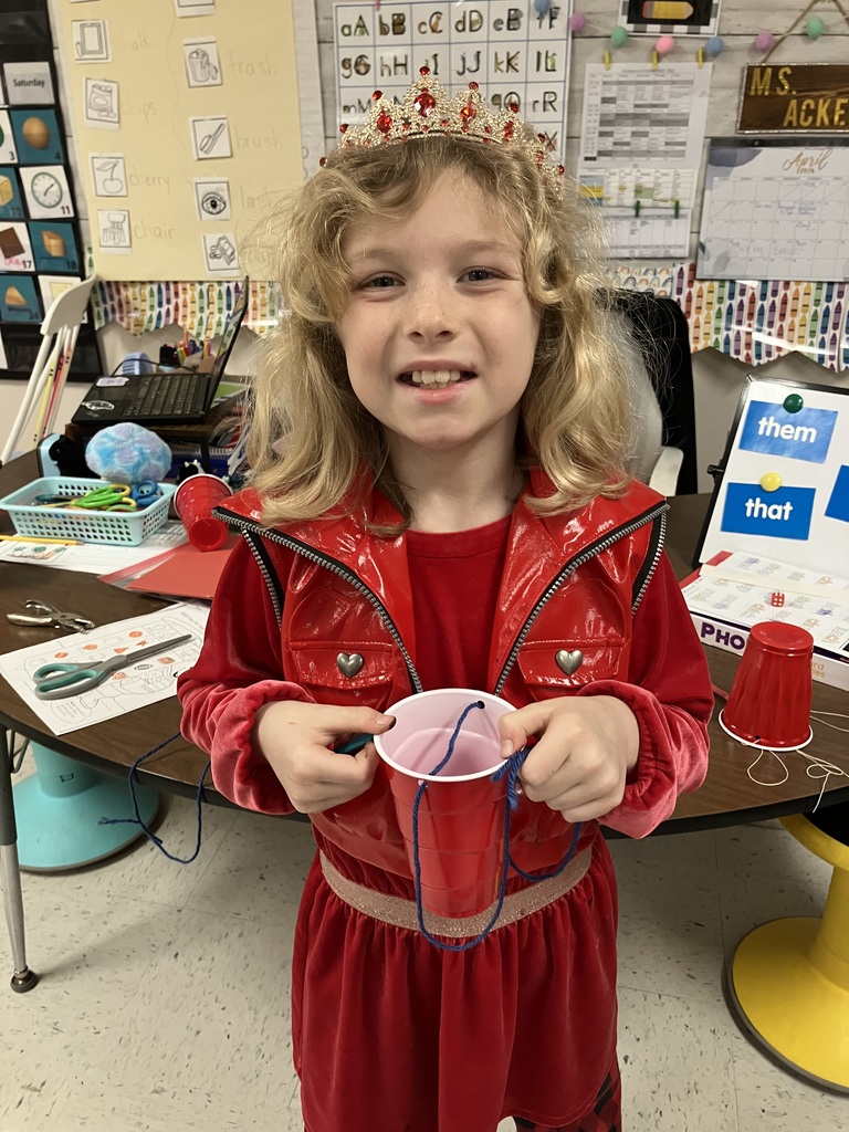  A close-up of a young girl with blonde hair wearing a gold and red tiara and a shiny red vest. She is smiling broadly while holding a red plastic cup with blue yarn attached to it.  I